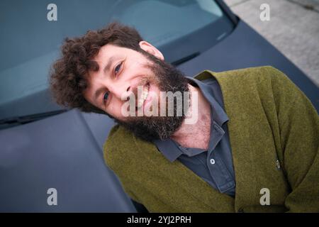 Homme barbu souriant dans une veste verte appuyée décontractée contre une voiture. Banque D'Images