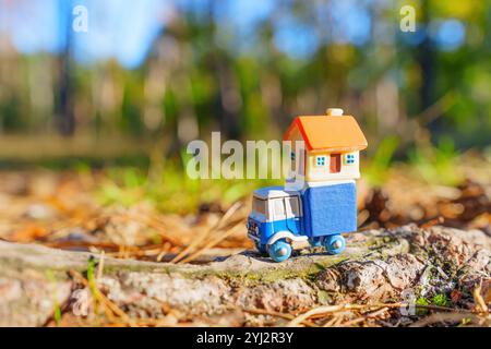 Petit camion-jouet transportant une maison miniature, positionné sur une bûche avec un fond extérieur vibrant avec des arbres et la lumière du soleil. Banque D'Images