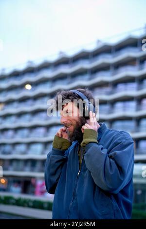 Homme souriant en veste bleue appréciant la musique sur les écouteurs à l'extérieur avec un immeuble d'appartements en arrière-plan au crépuscule, Belgique Banque D'Images