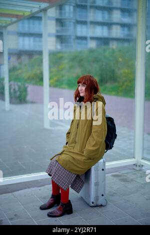 Jeune femme rousse assise sur une valise argentée à un arrêt de bus, portant un manteau vert, une jupe à carreaux et des bottes rouges, Belgique Banque D'Images