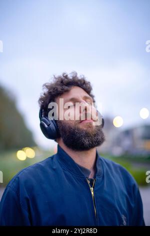 Homme barbu appréciant la musique avec les yeux fermés portant des écouteurs noirs à l'extérieur au crépuscule. Banque D'Images