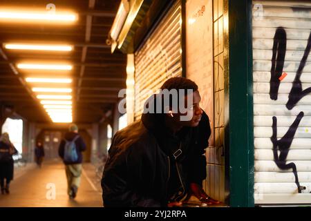 Un jeune homme regarde autour du coin, appuyé sur une fenêtre de service à volets, avec un éclairage chaleureux au-dessus et des passants au loin, Berlin, Allemagne Banque D'Images