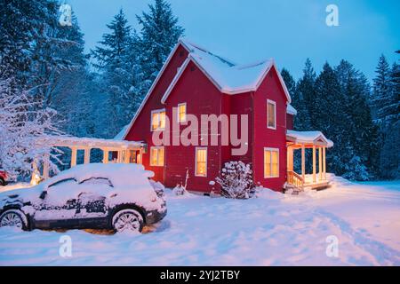 Maison rouge illuminée au crépuscule dans un paysage enneigé avec une voiture enneigée garée devant. Banque D'Images