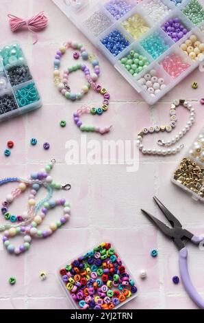 Des fournitures d'artisanat, y compris des perles colorées, des perles de lettre, de la ficelle, des fermoirs et des pinces, sont disposées sur une surface de tuile rose, prête pour la fabrication de bijoux Banque D'Images