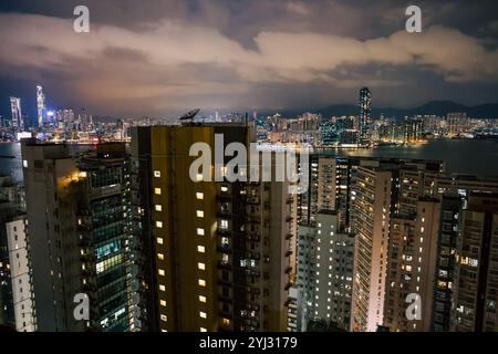 Le paysage urbain présente des bâtiments imposants et des lumières vibrantes qui se reflètent sur l'eau, mettant en valeur Hong Kong la nuit sous un ciel nuageux. Banque D'Images