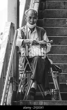 Un homme âgé est assis sur un escalier à Hong Kong, plongé dans la lecture d'un journal, mettant en scène un moment de calme au milieu de l'agitation urbaine. Banque D'Images