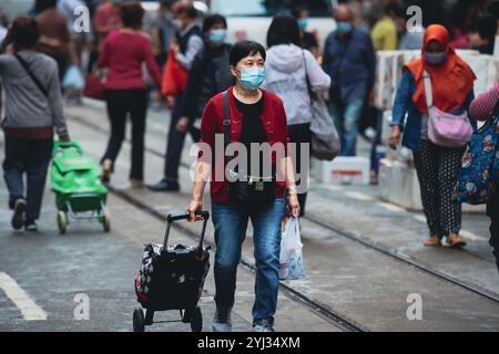 Une femme transportant des sacs et poussant un chariot traverse un marché de rue animé dans le centre de Hong Kong, entourée de piétons. Banque D'Images