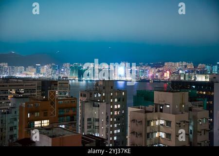 La ligne d'horizon de Hong Kong scintille avec les lumières vibrantes des bâtiments, se reflétant sur le port alors que le soleil se couche derrière les montagnes. Banque D'Images