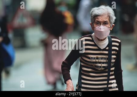 Une femme âgée navigue dans les rues animées de Hong Kong tout en portant un masque, mettant en valeur la vie quotidienne dans un cadre urbain dynamique. Banque D'Images