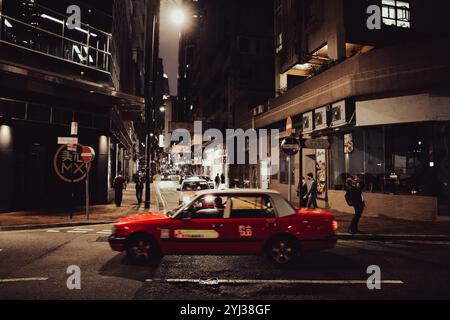 Un taxi rouge navigue dans une rue tranquille de Hong Kong illuminée par les lumières de la ville après le coucher du soleil. Banque D'Images