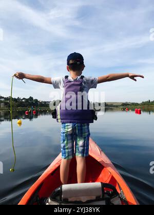 Un jeune garçon portant un gilet de sauvetage se tient sur un kayak les bras tendus, profitant d'une journée ensoleillée sur un lac serein. Banque D'Images