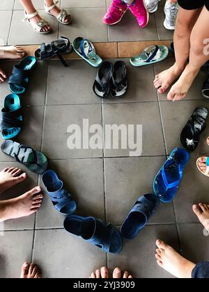 Une variété de tongs colorées et de sandales sont disposées en forme de coeur sur un sol carrelé, entouré par les pieds des enfants, transmettant un sens de commu Banque D'Images