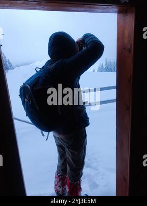 Une personne se tient dans une porte de cabine, photographiant le paysage enneigé. Ils portent des vêtements d'hiver et un sac à dos, mettant en valeur l'aventure et l'Explor en plein air Banque D'Images