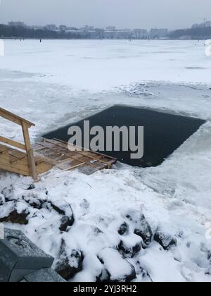 Un lac gelé avec un trou coupé pour la baignade hivernale, entouré de neige et de rochers. L'image montre une plate-forme en bois menant à l'eau glacée, sous un g. Banque D'Images