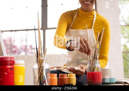 Femme peignant à la maison, souriant tout en sélectionnant des pinceaux de pot Banque D'Images