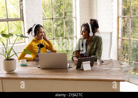 Deux amies féminines multiraciales enregistrant un podcast à la maison, souriant et portant des écouteurs Banque D'Images