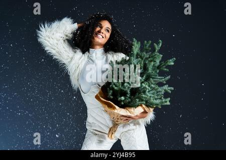 Une belle femme embrasse la magie hivernale tout en tenant joyeusement un petit sapin de Noël, habillé avec élégance. Banque D'Images