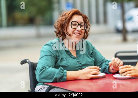 Femme handicapée en fauteuil roulant électrique buvant du café avec un ami assis dans une cafétéria extérieure dans la ville Banque D'Images