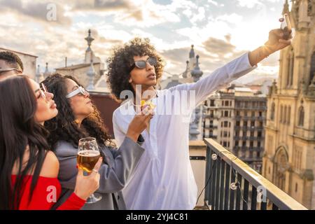 Jeune homme prenant selfie avec deux femmes de beauté dans la partie sur le toit avec vue sur la ville pendant le coucher du soleil Banque D'Images