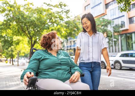 Femme heureuse caucasienne handicapée utilisant un fauteuil roulant parlant tout en marchant avec un ami asiatique le long de la ville Banque D'Images