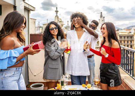 Groupe multiracial d'amis couvrant les yeux d'un homme lui donnant un cadeau dans une fête sur le toit Banque D'Images