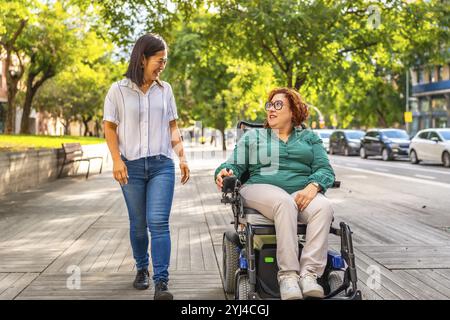 Femme handicapée utilisant un fauteuil roulant électrique et un ami chinois parlant tout en marchant le long de la ville Banque D'Images