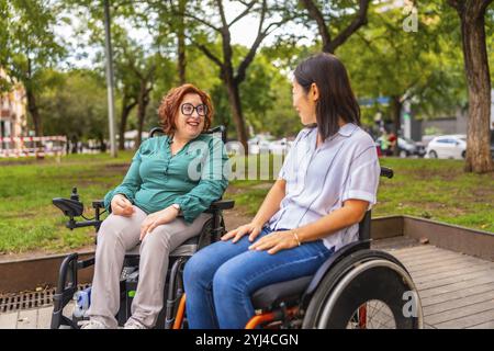 Femmes handicapées multiethniques assises sur des fauteuils roulants discutant ensemble dans un parc de la ville Banque D'Images