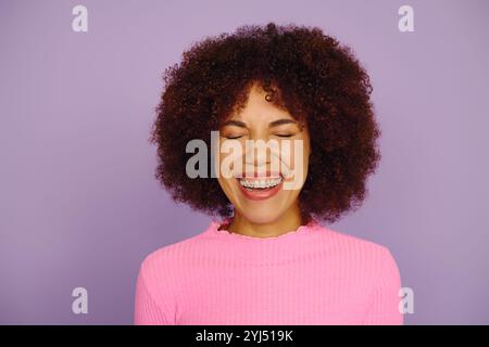 Une jeune femme afro-américaine aux cheveux bouclés rayonne de joie, souriant chaleureusement sur un fond violet vibrant. Banque D'Images