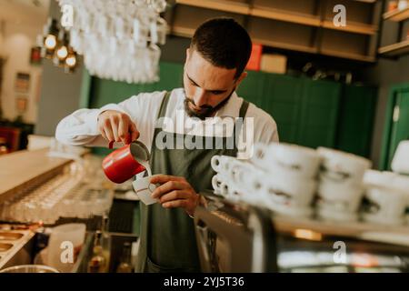 Dans un café chaleureux et accueillant, un barista dévoué verse habilement du lait dans une tasse à café, créant une expérience délicieuse pour les clients. Banque D'Images