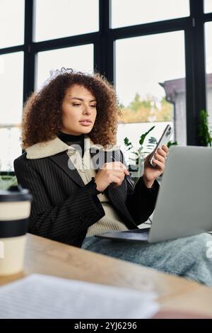 Une femme concentrée sur ses tâches utilise un ordinateur portable et un téléphone dans un espace de travail ensoleillé. Banque D'Images