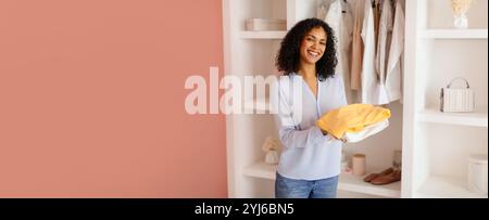 Une femme souriante se tient dans une pièce élégante, tenant des vêtements pliés de couleurs vives et entourée d'articles soigneusement disposés sur des étagères, reflétant un JO Banque D'Images