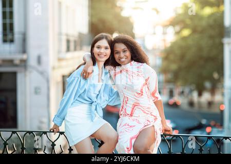 Deux jeunes femmes sourient et posent ensemble sur une balustrade dans un environnement urbain animé, profitant de la chaleur du soleil et de leur temps en plein air. Banque D'Images