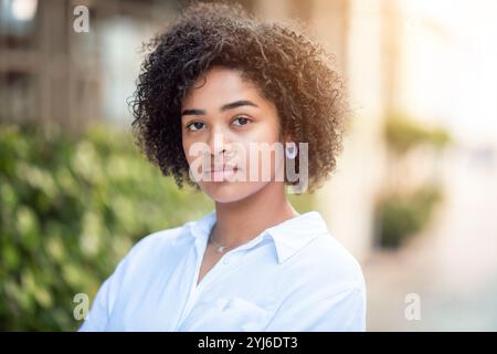 Une jeune femme se tient debout avec un comportement détendu, mettant en valeur ses cheveux bouclés tout en portant une chemise blanche. L'arrière-plan présente de la verdure, créant une ambiance chaleureuse Banque D'Images