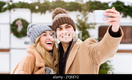 Un couple heureux partage un moment alors qu’ils prennent un selfie en vêtements d’hiver, entouré de neige et de décorations festives. Leurs expressions reflètent la joie et Banque D'Images