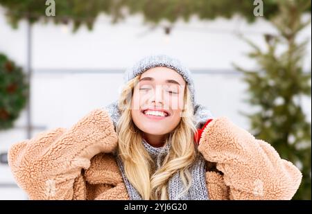 Une jeune femme profite d'une belle journée d'hiver. Elle porte un manteau de fourrure confortable et un chapeau en tricot, souriant largement avec ses yeux fermés, entourée de neige et Banque D'Images