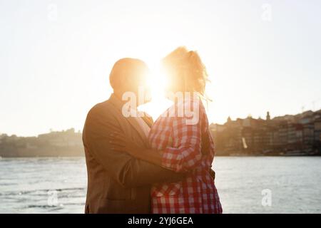 Un couple partage un moment tendre alors que le soleil se couche derrière eux, illuminant leurs silhouettes contre le front de mer tranquille. La scène capture Love A. Banque D'Images