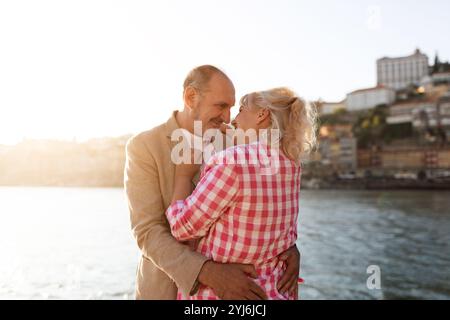Couple mûr embrasse avec amour près de la rivière comme le soleil se couche, jetant une lueur chaude sur leurs visages joyeux, entouré de vues panoramiques de la ville en t Banque D'Images