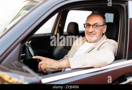 Un homme confiant avec des lunettes est assis dans le siège du conducteur d'une voiture, pointant vers l'extérieur pendant qu'il regarde la caméra. La lumière chaude du jour met en valeur son rel Banque D'Images