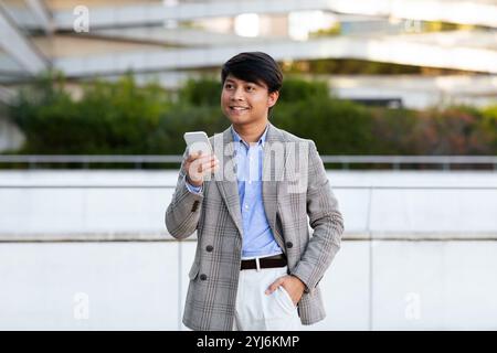 Un homme asiatique bien habillé sourit en regardant son smartphone tout en se tenant dehors parmi des bâtiments contemporains. Le réglage suggère un casual yet p Banque D'Images