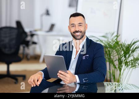 Un homme confiant vêtu d'un élégant costume rayé est assis sur un bureau dans un bureau contemporain. Il sourit tout en tenant une tablette, entouré d'un propre, ou Banque D'Images