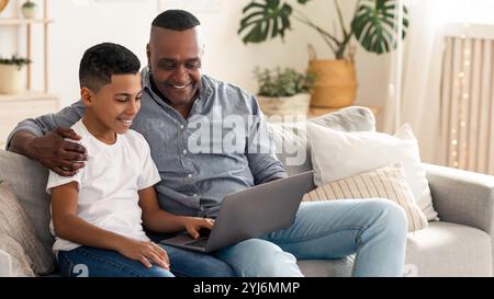 Grand-père noir soignant enseignant à son petit-fils preteen comment utiliser l'ordinateur, assis avec l'ordinateur portable sur le canapé dans le salon, espace libre Banque D'Images