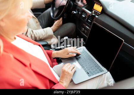 Une femme dans une veste rouge tape sur son ordinateur portable alors qu'elle est assise dans le siège passager d'une voiture. Le conducteur, partiellement visible, est concentré sur la route, showc Banque D'Images