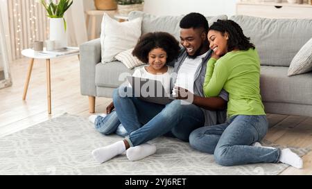 Planifier des vacances ensemble. Papa noir, maman et petite fille utilisant un ordinateur portable à la maison, assis sur le sol dans le salon, espace de copie Banque D'Images