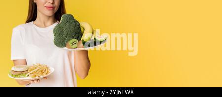 Sain vs malsain. Dame méconnaissable choisissant entre des assiettes avec des fruits et légumes biologiques et de la malbouffe, fond jaune, recadré Banque D'Images