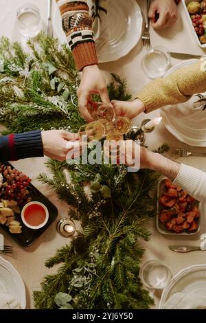 Photo descendante d'amis méconnaissables assis à la table décorée pour Noël et serrant des verres en cristal remplis de vin mousseux Banque D'Images
