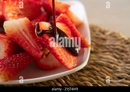 Gros plan de chocolat qui s'égoutte sur une fraise sur une assiette carrée blanche avec des fraises hachées sur une table en osier Banque D'Images