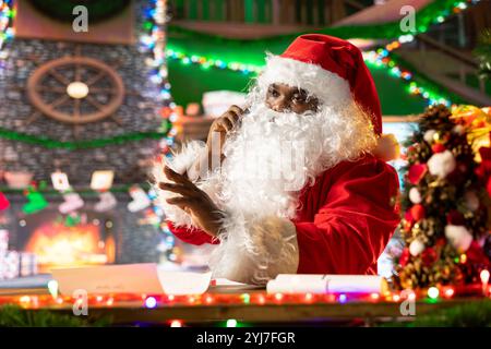 Père Noël noir répondant à un appel téléphonique dans son atelier du pôle Nord, préparant la fête de la veille de Noël. Homme barbu blanc avec costume rouge soulignant la tradition de vacances. Banque D'Images