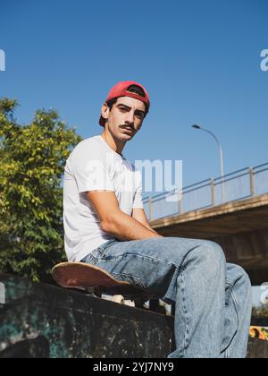 Jeune homme en vêtements décontractés assis sur le skateboard à l'extérieur Banque D'Images