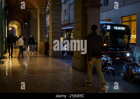 Bologne, Italie. 8 octobre 2024 - les gens marchent sous des arches historiques avec un bus dans la rue dans la soirée Banque D'Images
