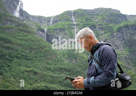 Un homme aux cheveux gris dans les années 40 avec une expression focalisée tient une télécommande de drone, debout avec les cascades de Kjelfossen en arrière-plan près de Gudva Banque D'Images